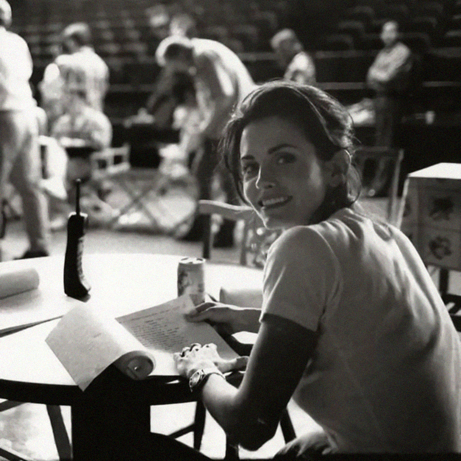 A woman smiles at the camera while sitting at a round table, holding a pen over papers. A can and a walkie-talkie are nearby. Blurred people and chairs fill the background, suggesting a busy, casual work setting.