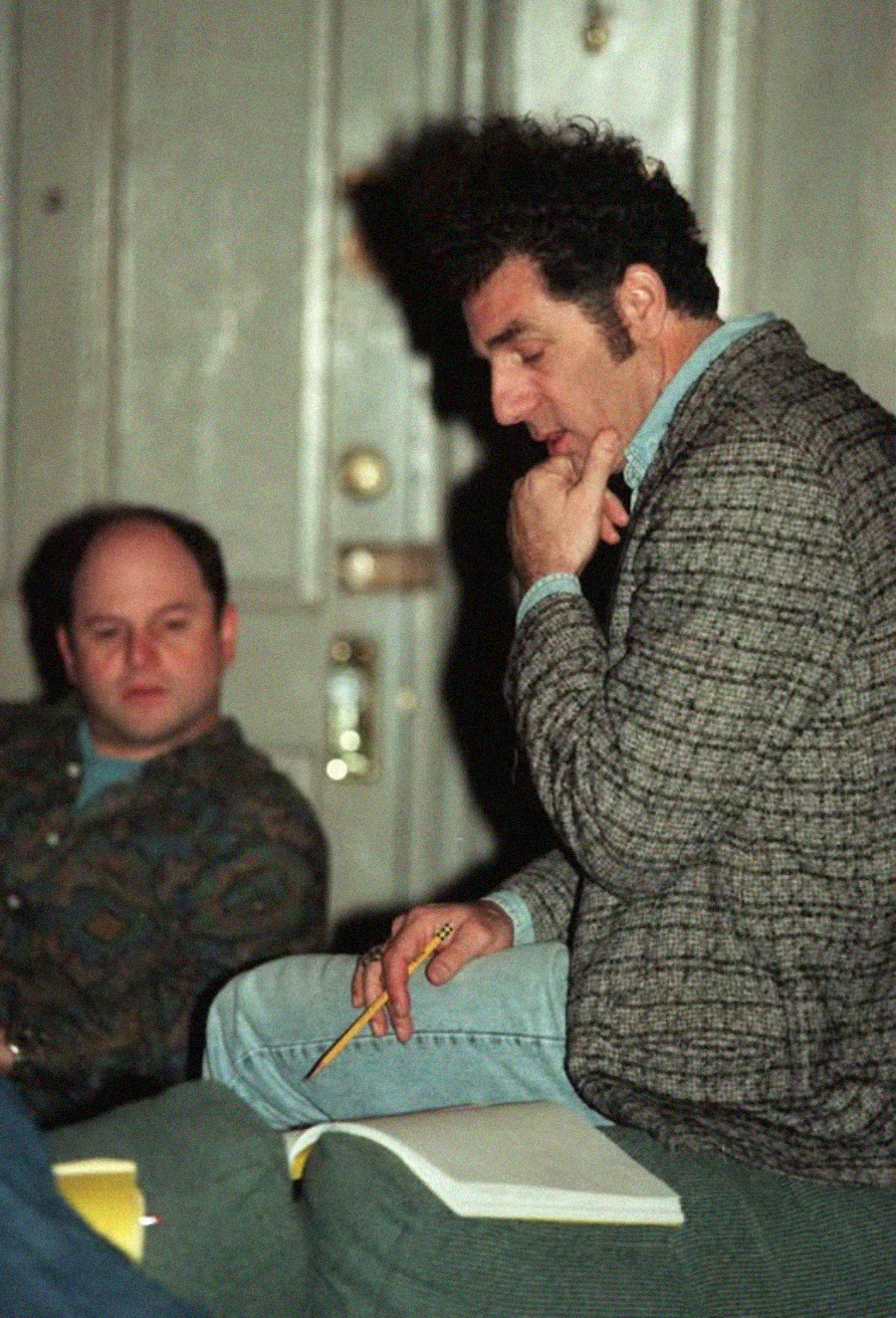 Two men sit indoors; one is holding a pencil and notebook, appearing deep in thought, while the other looks on. The background features a closed door and muted lighting, suggesting a contemplative or serious setting.