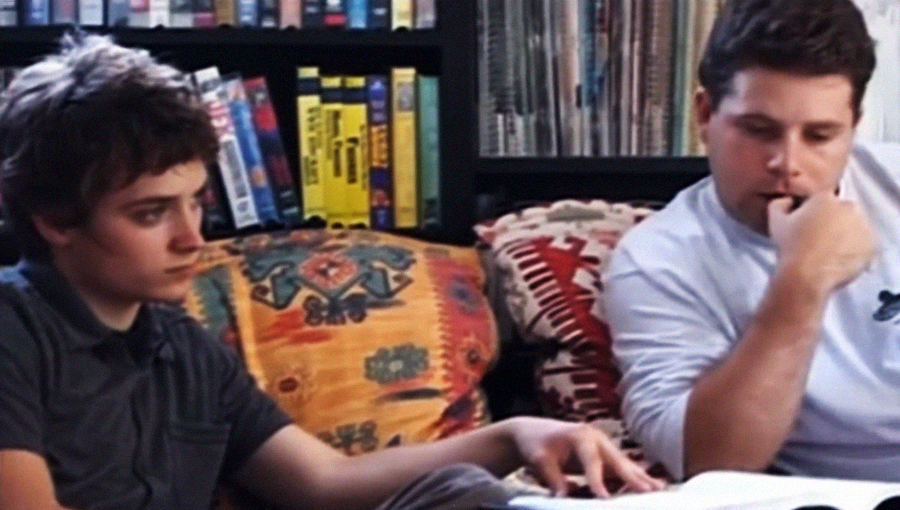 Two people sit on a couch with colorful cushions, looking at an open book together. Shelves filled with books and VHS tapes are in the background. Both appear focused and engaged in the material.