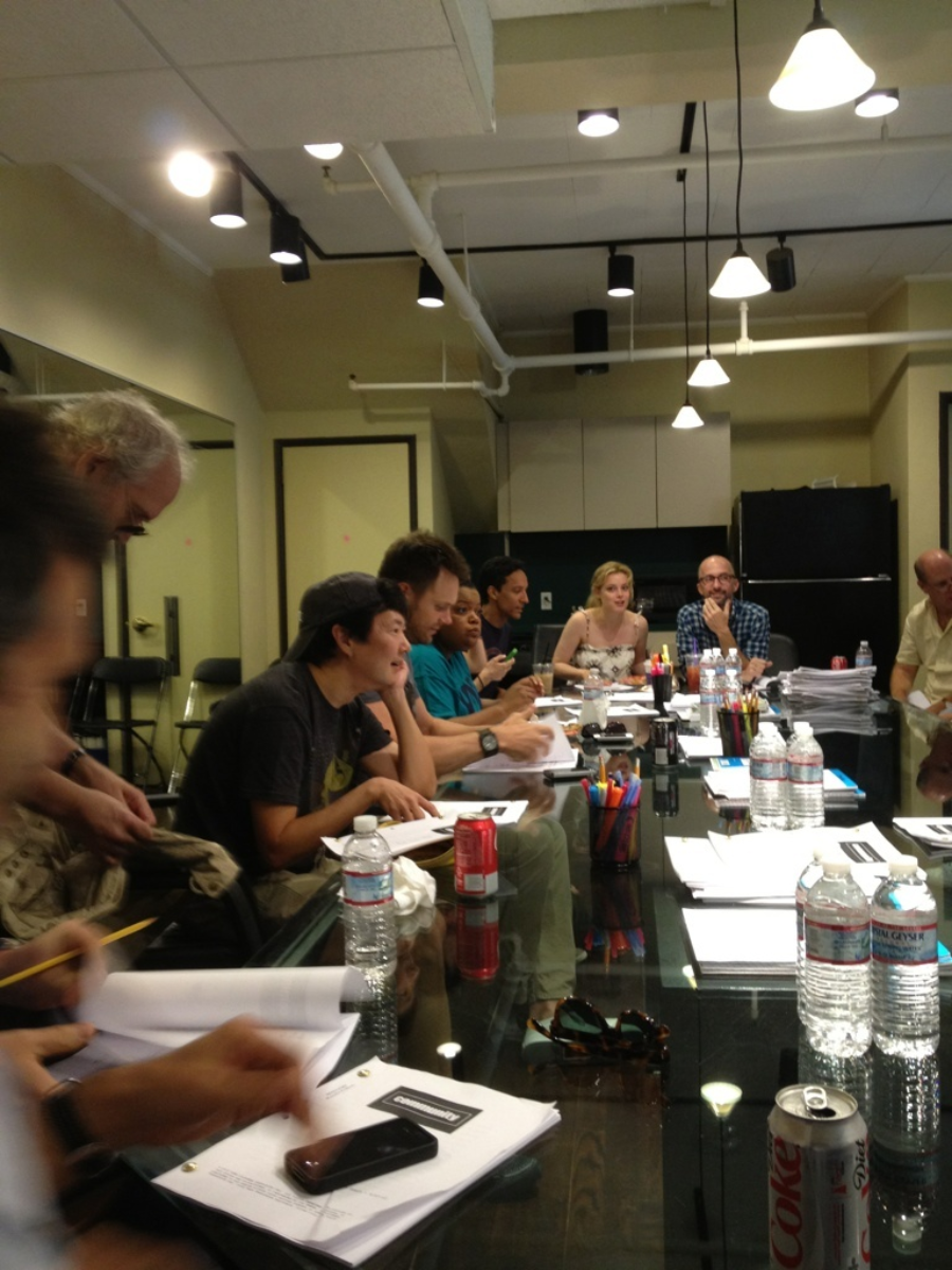 A group of people sit around a glass table with scripts, drinks, and stationery, engaged in discussion in a well-lit meeting room with overhead lights and exposed pipes.