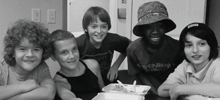 Five smiling children sit around a table with takeout food containers, posing for a casual black-and-white photo. Some are leaning on the table, and one wears a bucket hat. They appear happy and relaxed.