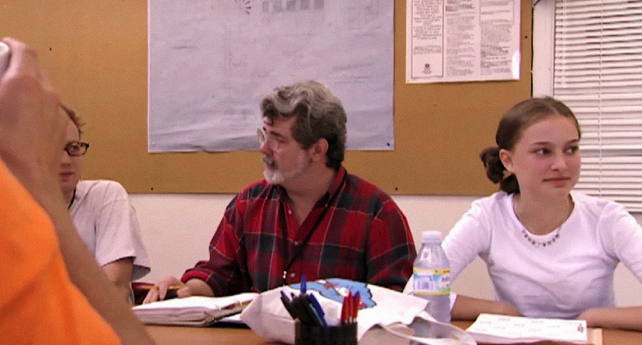 Three people sit at a table with notebooks and pens, engaged in discussion. The man in the center wears a red plaid shirt, and a young woman in a white shirt sits to his right. A corkboard is on the wall behind them.