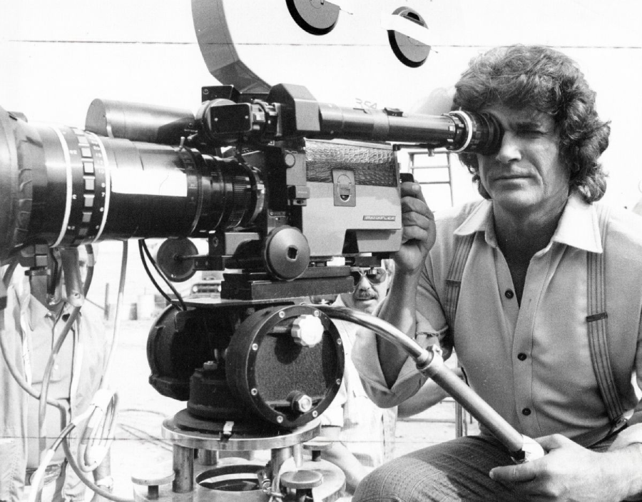 A man with curly hair and sideburns looks through the viewfinder of a large vintage film camera on set, with technical equipment visible in the background.