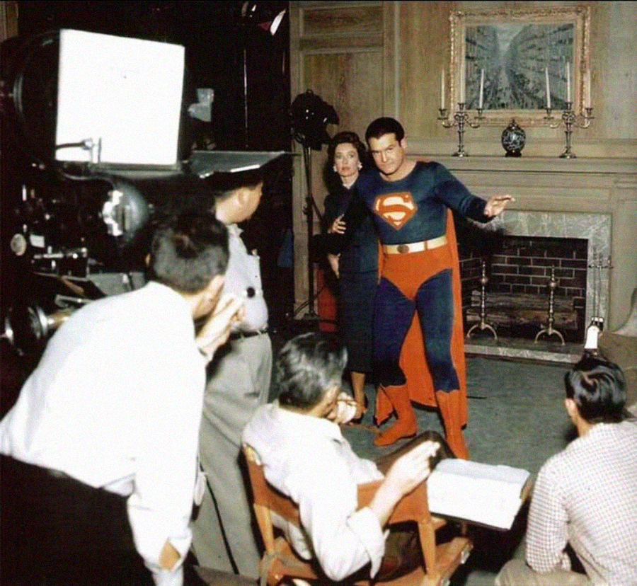 A man dressed as Superman poses on a vintage film set, surrounded by crew members and a woman in a dark dress. A large camera and director's chair are visible in front of a fireplace with ornate decorations.