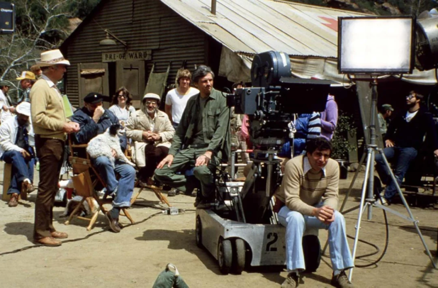 A film crew is shooting a scene outdoors on a dirt lot with equipment and large lights. Several people sit and stand around, while one operates a camera mounted on a dolly track. A rustic building labeled "PAD-OF-WARD" is in the background.