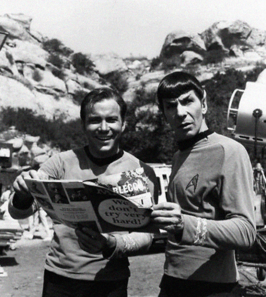 Two men in Star Trek uniforms stand outdoors, holding and reading a magazine together. One smiles at the camera while the other looks surprised. Rocky hills and filming equipment are visible in the background.