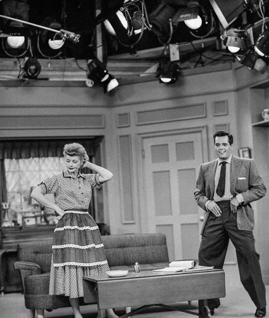 A woman in a polka dot dress and a man in a suit stand in a living room set under studio lights, both striking playful poses, on a black-and-white TV show set.