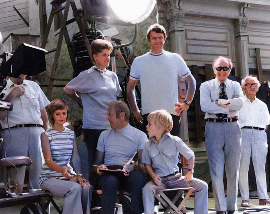 A group of men, a woman, and a boy are gathered on a film set outdoors, surrounded by lighting equipment. Some are standing, others are seated, and all are casually dressed, appearing relaxed during a break in filming.