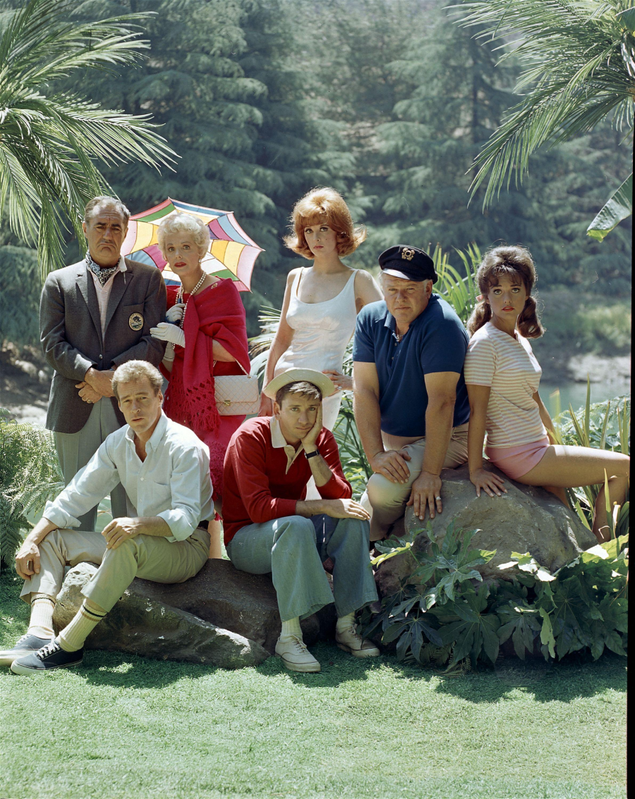Seven people pose outdoors in a lush, green setting with palm trees. Some are sitting on rocks while others stand behind them; one woman holds a colorful umbrella. They wear casual and vintage-style clothing.