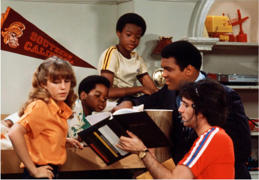 A group of four children and an adult man gather around a book in a bedroom decorated with a "Southern California" pennant and model airplane; they appear to be discussing or studying together.