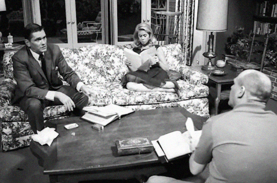 Three people are in a living room: one man in a suit sits on a floral sofa, a woman reads a magazine with her legs up, and a bald man sits at a coffee table covered with books and papers.