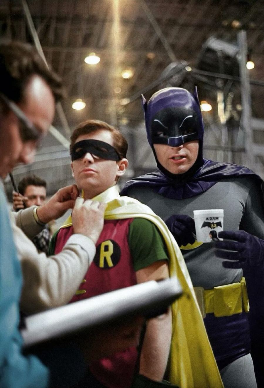 Two actors in classic Batman and Robin costumes stand on a set. Robin is having his cape adjusted by a crew member, while Batman holds a coffee cup and looks toward the camera. The background shows studio lights and equipment.