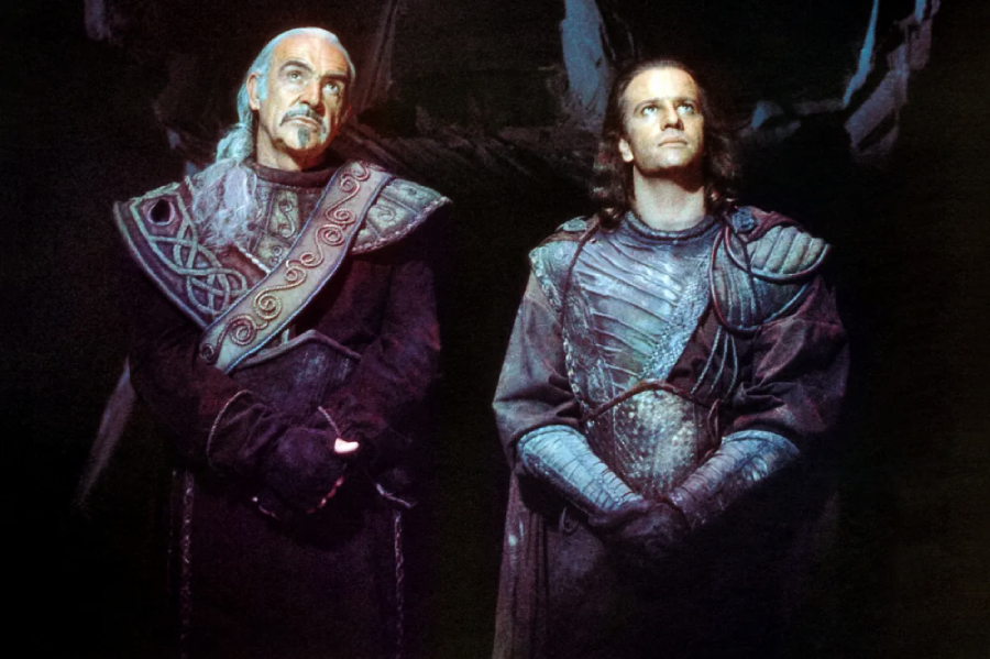 Two men in ornate, medieval-style armor stand side by side in a dimly lit setting, looking upward. The man on the left has a beard and white hair, while the man on the right has long brown hair.