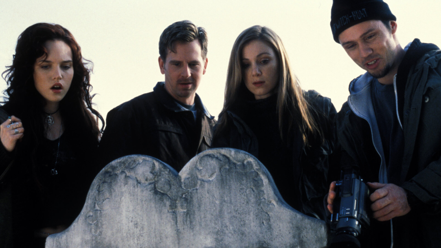Four people stand closely together outdoors, looking down at an old, weathered gravestone. One person holds a video camera, and their expressions are serious and curious.
