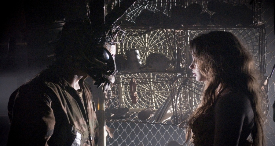 A man wearing a dark, animalistic mask faces a woman with long, messy hair in a dimly lit room filled with wire cages and cluttered shelves. Shadows enhance the scene’s tense, mysterious atmosphere.