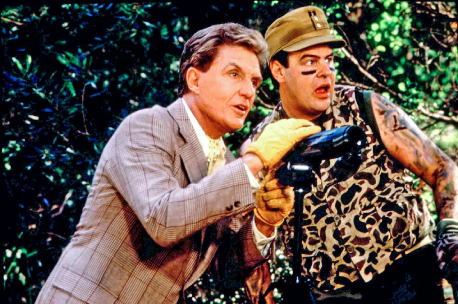 Two men stand outdoors surrounded by greenery; one wears a plaid suit and yellow gloves, holding a black object, while the other wears a camo outfit and cap with black face paint, looking in the same direction.
