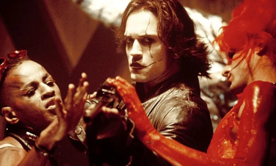 Three people in dramatic makeup and costumes stand close together. The central figure, with pale face paint and black clothing, stares intensely forward while two others, one with red body paint, interact with him. The scene is tense and theatrical.
