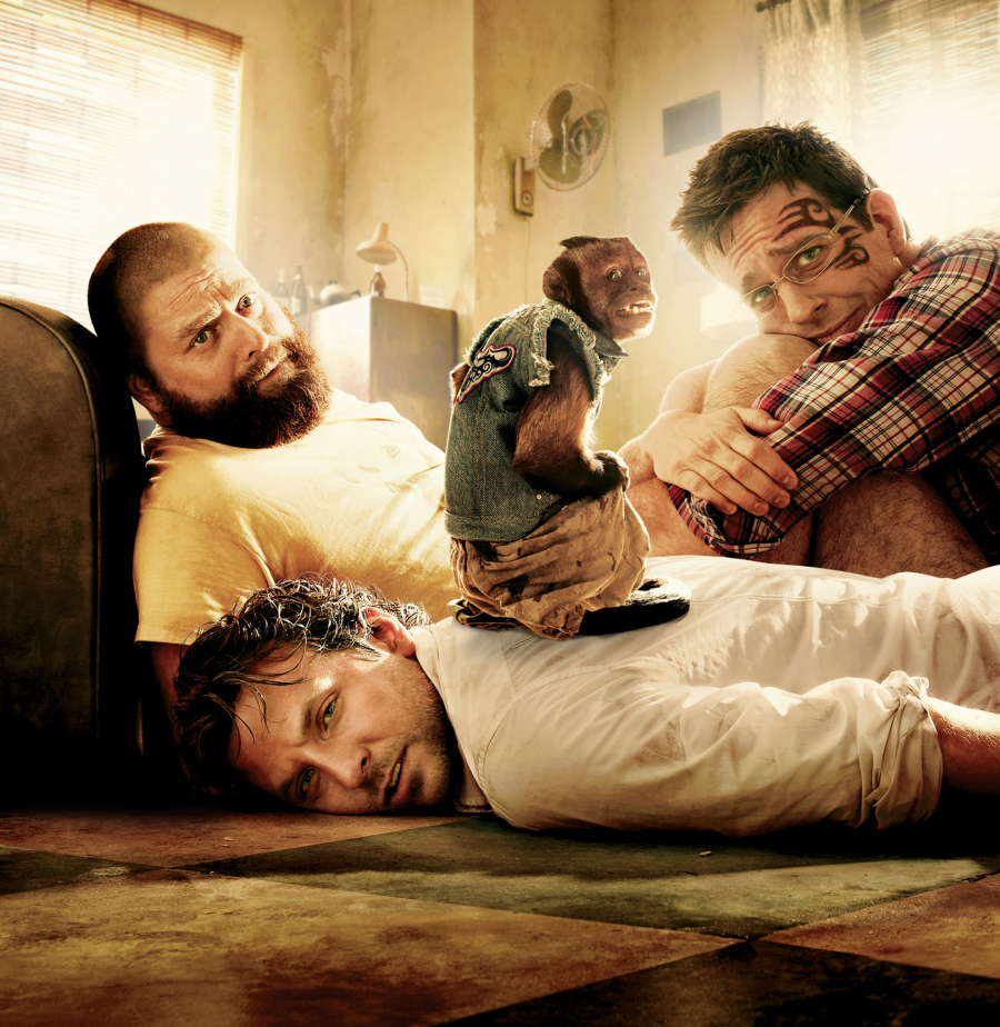 Three disheveled men sit and lie on a messy floor in a sunlit room; one has a face tattoo, another looks exhausted, and a small monkey in a denim vest stands on the back of the man lying down.
