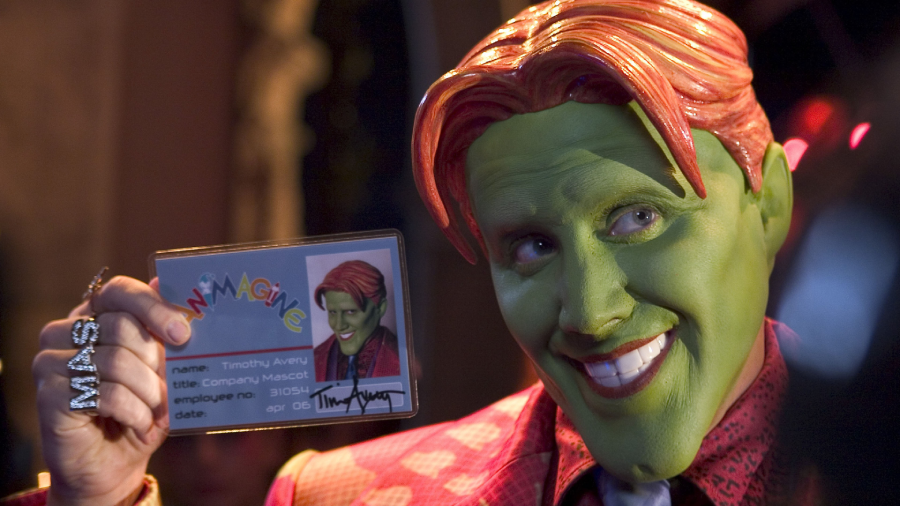 A person with green face paint, orange hair, and a red suit smiles while holding up an ID badge with their photo on it. The badge reads "IMAGINE" and the person's ring spells "MASK.