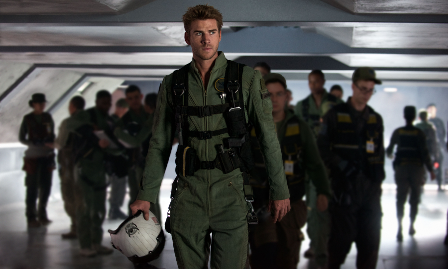 A man in a green flight suit and harness walks confidently while holding a white helmet; several other uniformed people are seen blurred in the background inside a futuristic military facility.