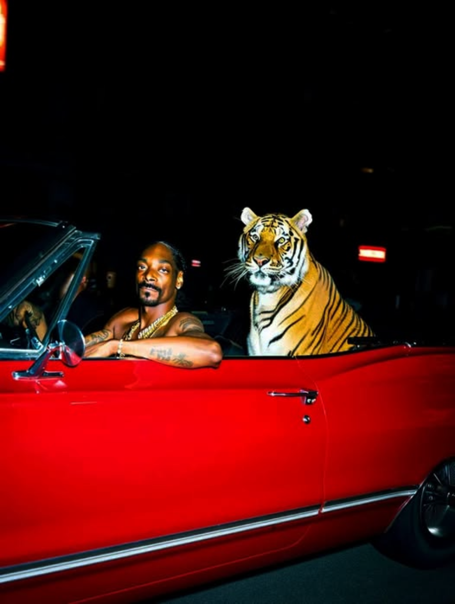 A man wearing gold chains sits in a red convertible at night, smiling at the camera. A tiger is seated beside him in the passenger seat, both illuminated against the dark background.