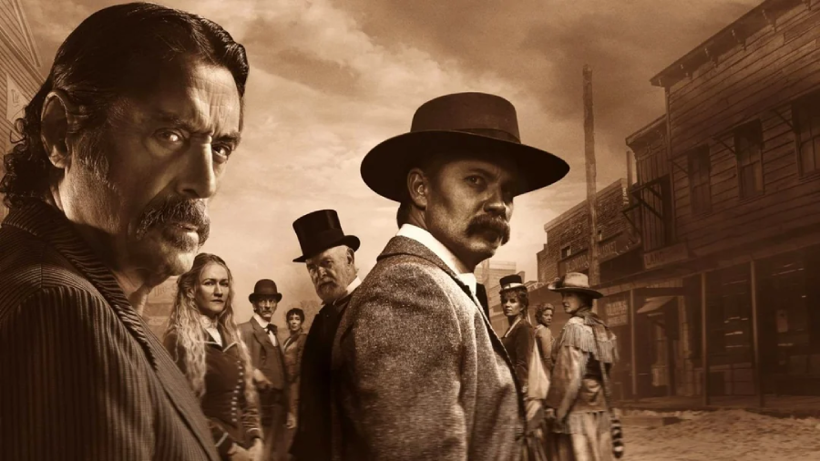 A group of people dressed in Old West attire stand in a dusty, sepia-toned frontier town; two men with serious expressions stand in the foreground, while others watch from behind.