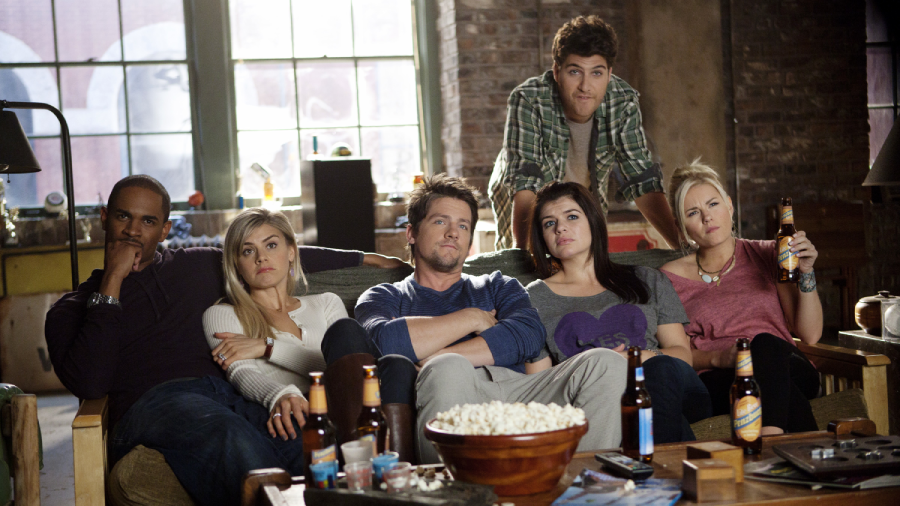 Six people sit closely on a couch and chairs in a cozy living room, watching something off-camera. There are drinks and snacks on the table in front of them. Everyone looks relaxed, with neutral or focused expressions.