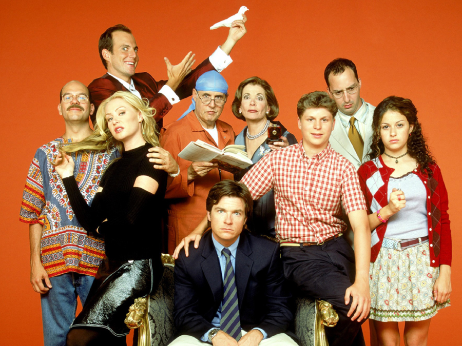 Nine people pose in front of a plain orange background, with varied expressions. Some stand while others sit. One man holds a white dove. The group appears quirky and diverse in style and attire.