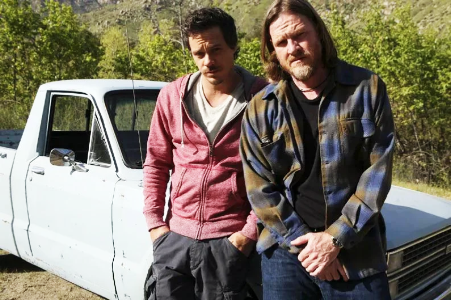 Two men stand in front of an old white pickup truck in an outdoor setting. One wears a red hoodie and gray pants; the other wears a plaid shirt and black t-shirt. Trees and hills are visible in the background.