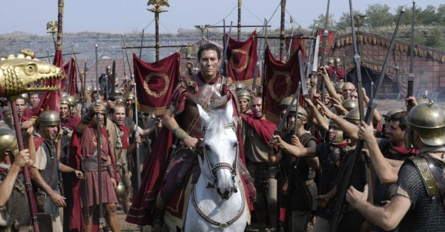A man in Roman armor rides a white horse, surrounded by Roman soldiers holding red banners and spears, with a golden dragon-headed standard visible. The scene appears set in ancient times, possibly during a military campaign.
