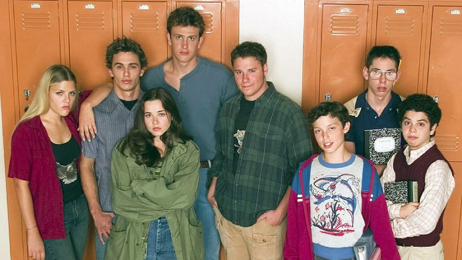 Eight teenagers stand in front of orange school lockers, posing for a group photo. They are dressed in casual 1980s-style clothing, with varied expressions, some smiling and others serious.