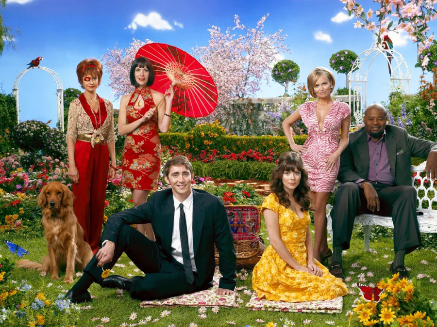 Six people and a golden retriever pose in a colorful, flower-filled garden. Two women stand with a red parasol, one woman sits in a yellow dress, and a man in a suit sits beside her. The scene appears bright and whimsical.