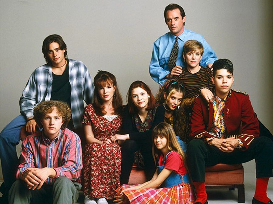 A group of eight people, including adults, teens, and children, pose together in casual and colorful clothing against a plain backdrop. Some are seated while others stand behind them, all looking toward the camera.