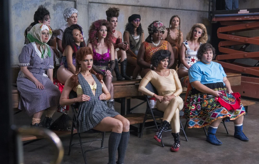 A group of women in colorful, dramatic costumes sit on benches and chairs in an industrial room, watching something off-camera with serious or focused expressions.