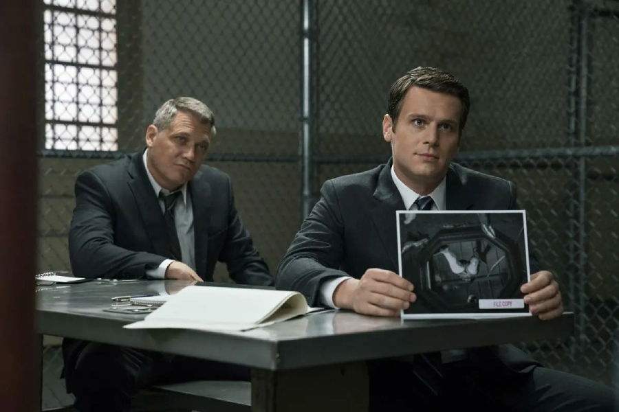 Two men in suits sit at a table in an interrogation room. One man holds up a black-and-white photo, while the other looks on. Papers are spread on the table, and a chain-link fence is visible in the background.