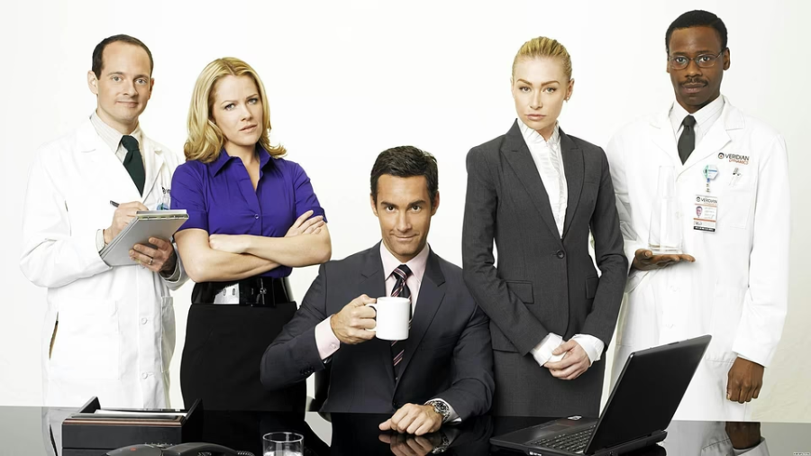 Five people stand together against a white background: two in white lab coats, two in business attire, and one man in a suit sitting at a desk holding a coffee mug, with a laptop in front of him.
