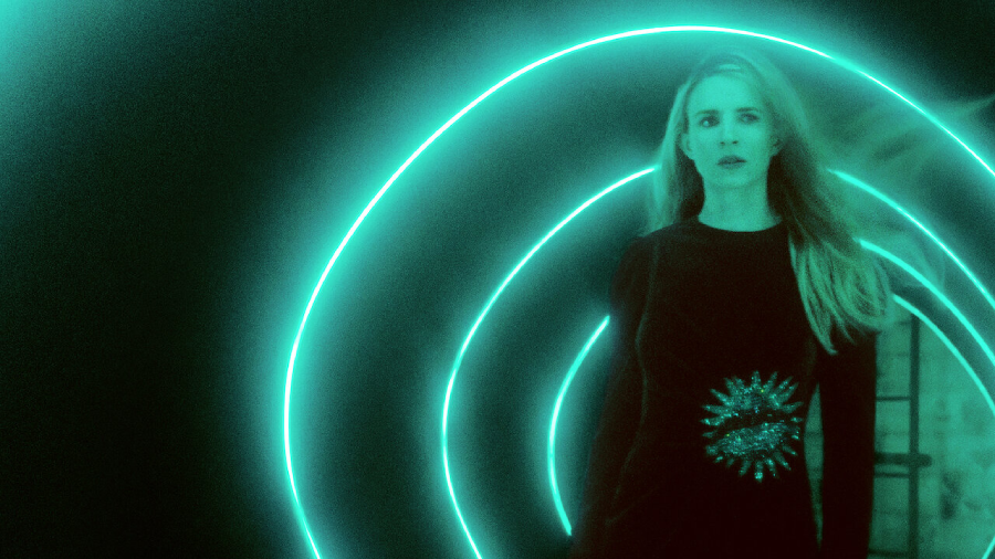 A woman with long blonde hair stands in a dark area, illuminated by bright, glowing teal light trails curving around her. She wears a black outfit with a sparkling sunburst design on the front.