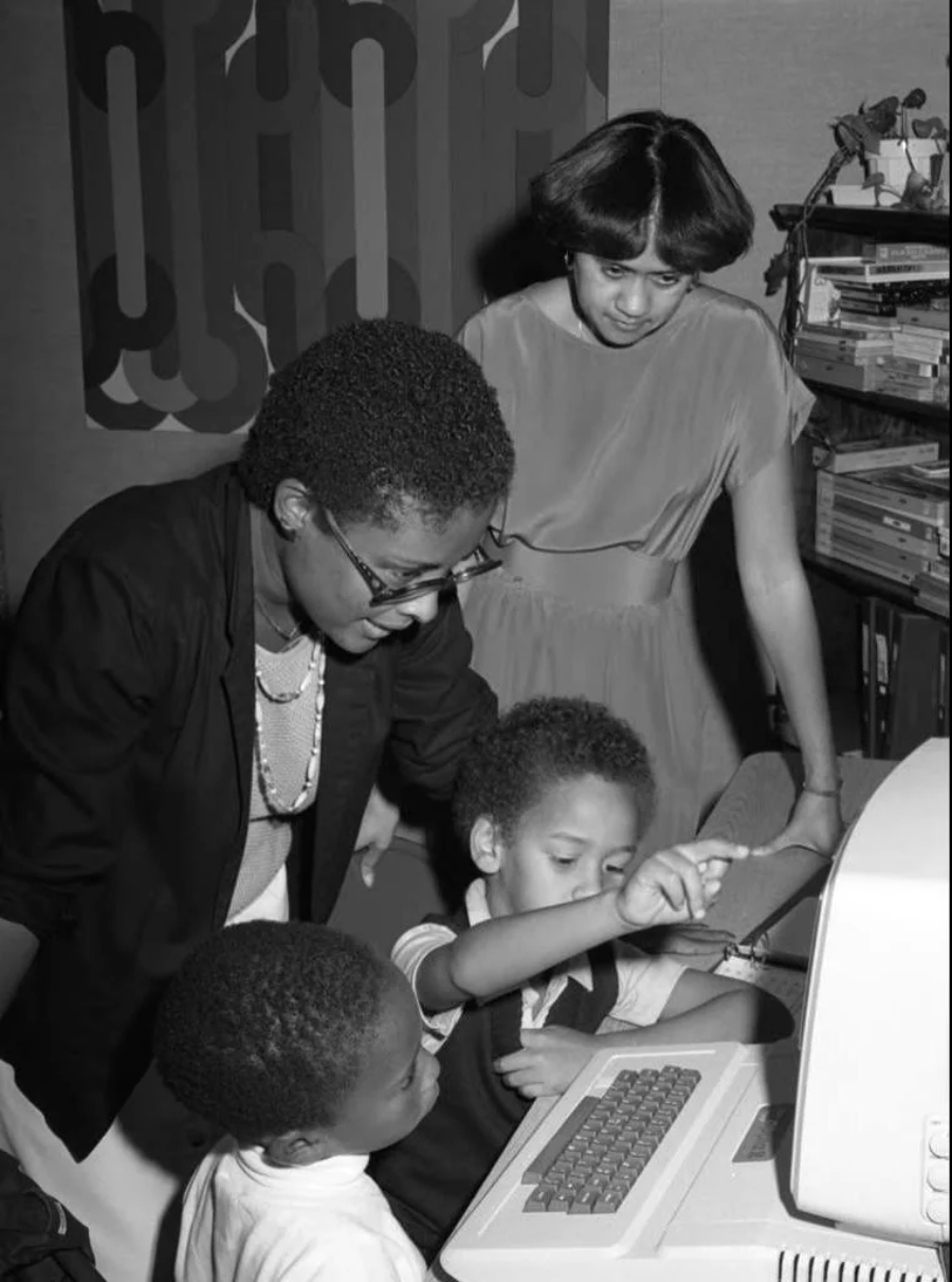 Two women and two young children stand around a vintage computer. One child points at the computer screen while the others watch with interest. The scene appears to be from the 1980s, in a home or classroom setting.
