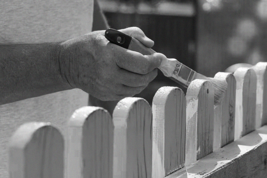 A person’s hand holds a paintbrush, painting the wooden slats of a picket fence outside. The image is in black and white, focusing on the hand and the fence.