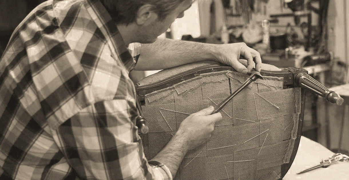 A person in a plaid shirt repairs the backrest of an upholstered chair using a tool in a workshop, with various tools and materials in the background.
