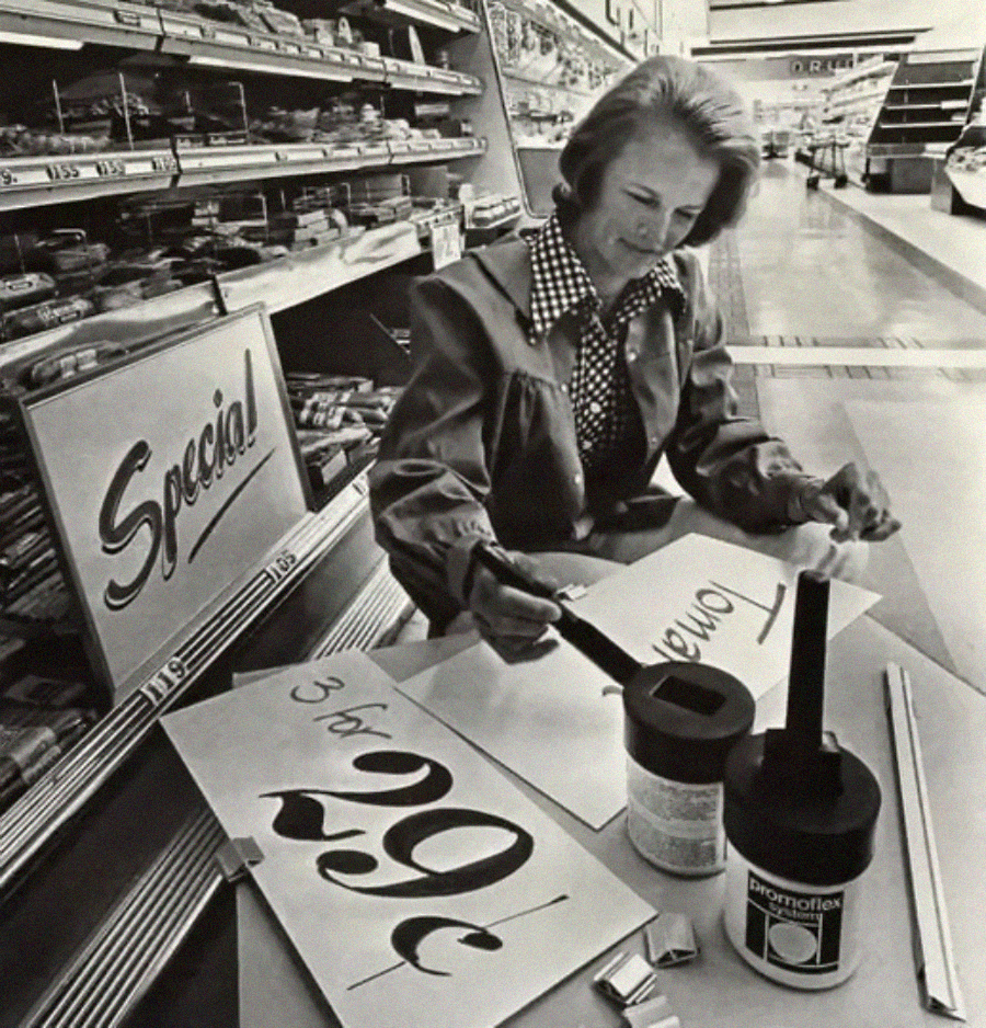 A woman sits in a grocery store aisle hand-painting sale signs, including one reading “3 for 29¢.” Paint pots and brushes are on the table, and shelves of food products line the background.