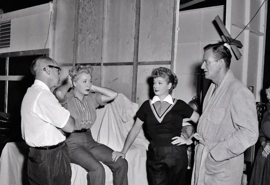 Four people talk on a film or TV set. Two women sit and stand in the middle; one looks surprised with hands near her head. Two men stand on either side, one in glasses, the other in a bathrobe. A large wooden wall is behind them.