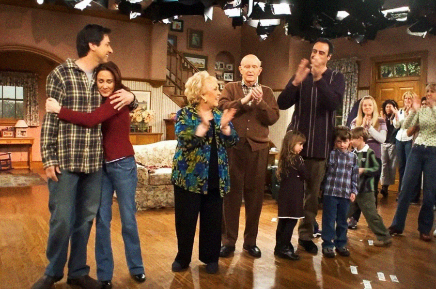 A group of people stand on a cozy living room set, smiling and applauding. Some hug, while others clap, and several children are present. Studio lights and crew are visible above, indicating a TV show or filming set.
