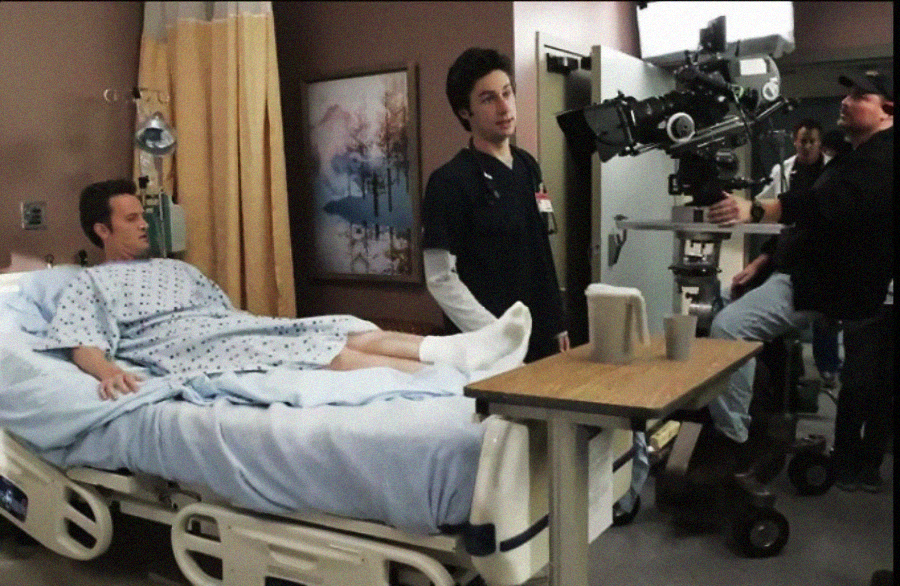 A hospital scene is being filmed; a patient lies in bed while an actor in scrubs stands nearby. A camera crew with filming equipment is positioned beside them, capturing the scene.