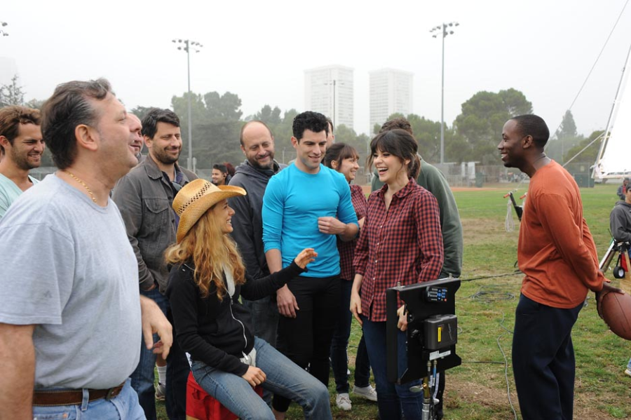 A group of people gather outdoors on a grassy field, laughing and talking. Some are standing, one person sits on a chair, and filming equipment is visible, suggesting a relaxed moment on a movie or TV set.