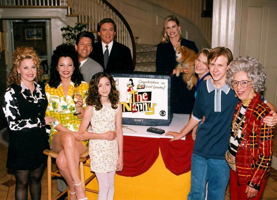 A group of people from "The Nanny" TV show cast pose around a TV displaying the show’s logo and a congratulatory message for their 100th episode, smiling in a warmly lit living room with a staircase in the background.