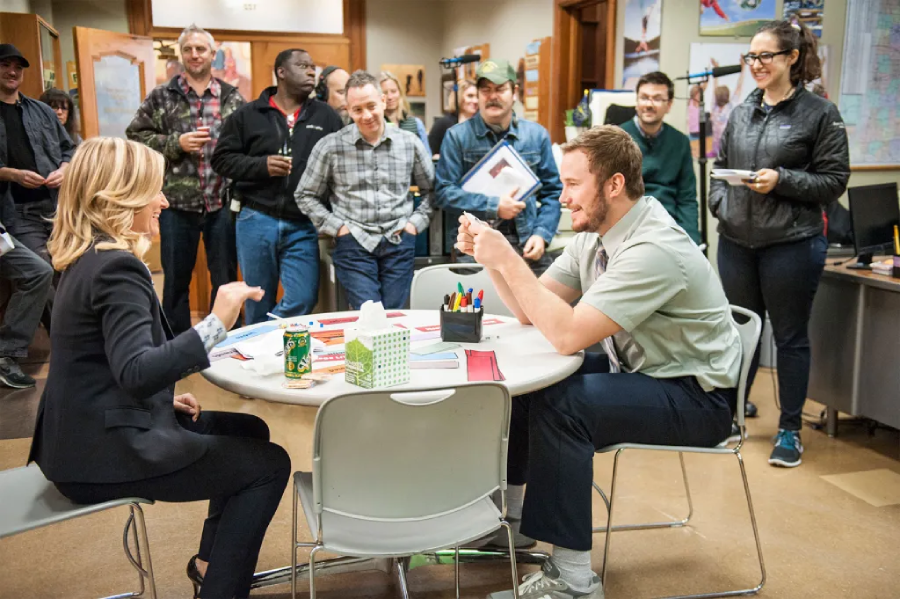 Two people sit across from each other at a round table, smiling and talking, while a group of people stand around them in an office setting, watching and laughing. The mood is lively and cheerful.