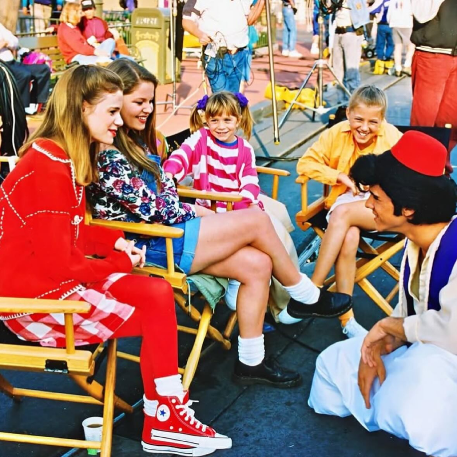 Five children, some in colorful outfits, sit on director’s chairs outdoors, smiling and laughing with a man dressed as Aladdin, wearing a red fez and white vest. People and film equipment are visible in the background.