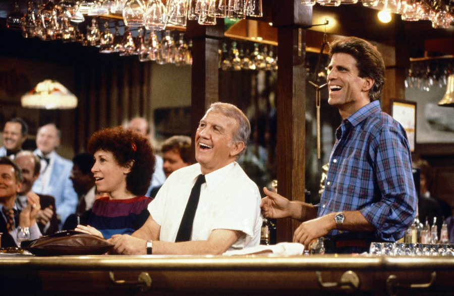 Three people stand behind a busy bar, laughing and smiling, with hanging glasses overhead and a group of patrons in the background watching them. The scene appears lively and cheerful.