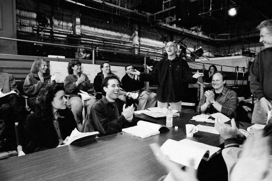 A black-and-white photo shows a group of people gathered around a table, laughing and clapping during what appears to be a script reading or rehearsal in a studio setting with papers and water bottles on the table.